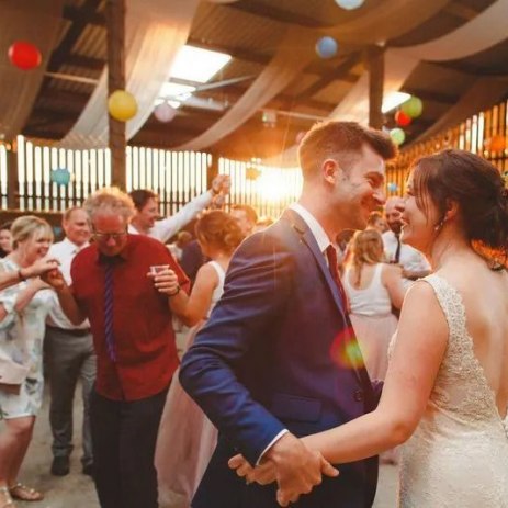 A couple dances at a lively wedding reception, surrounded by guests, colorful balloons, and string lights in a rustic venue.